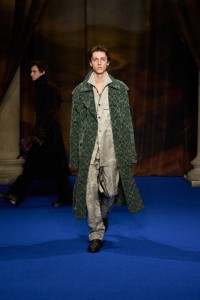 Saul Symon indossa un trench in misto viscosa con stampa barocca vine green, camicia con girasoli e pantaloni stile pigiama in seta jacquard taupe beige e stivali anchor grey. Il look è abbinato a un bracciale con pendente Knight placcato in argento e a un anello impilabile Hollow placcato in oro.