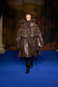 Edie Campbell indossa un parka con mantella in pelliccia sintetica taupe, imbottitura in nylon trapuntato stampato sherry red, felpa con cappuccio in misto lana mahogany red, pantaloni in cotone treacle brown e pebble beige. Abbinato a stivali alti fino alla coscia in nero e argento e bracciale Star Link placcato oro.