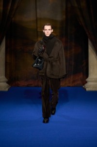 Jack Cooper indossa una giacca con collo sciallato in lana e cashmere bark mélange brown scuro, pullover a collo alto in misto lana walnut brown, pantaloni sartoriali in corduroy clove brown e borsa da bowling Highlands in pelle peat brown.