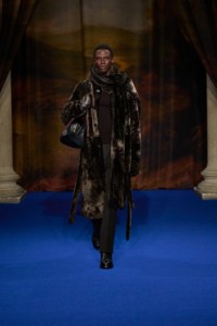 Cherif Douamba indossa un cappotto con sciarpa in shearling e un pullover con collo sciallato in lana a coste bark brown, camicia stile pigiama in seta Check rubus purple e pantaloni sartoriali raven brown.