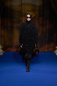Hejia Li indossa una mantella nera in misto lana mohair con frange all'uncinetto, un body in misto lana alpaca nera lavorato a nido d'ape e una gonna a pieghe in seta con girasoli nera, abbinati a stivali Cavalier in pelle truffle brown e borsa in pelle ink blue.