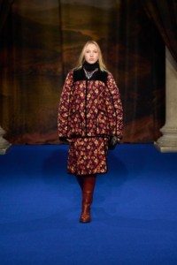 Lila Moss indossa una giacca con stampa a foglie e una gonna trapuntata poppy red, stivali Cavalier in pelle conker brown, orecchini a cerchio Charm placcati in oro e collana Star Bow smaltata in argento. 