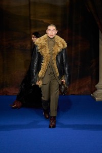 Sang Woo Kim indossa una giacca da aviatore in shearling nero, giacca mouliné e pantaloni tarmac grey e pebble beige, abbinati a un pullover a collo alto khaki brown e stivali in pelle mahogany red.